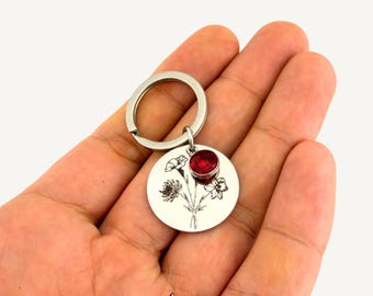May include: A circular silver-toned metal keychain featuring a black engraved floral bouquet design. A small, round, faceted red crystal charm is attached to the top of the disc, which hangs from a standard silver-toned metal split ring.