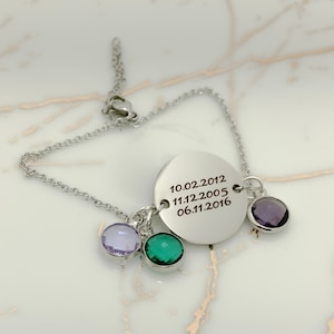 May include: A silver bracelet with a round charm engraved with the dates 10.02.2012, 11.12.2005, and 06.11.2016. The bracelet also has three small charms: one purple, one green, and one light purple.