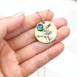 May include: Gold necklace with a round pendant featuring a holly sprig design and the name "Clara" engraved on it. A green gemstone is attached to the top of the pendant.