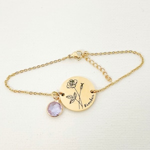 May include: Gold chain bracelet with a round charm featuring a rose and the name "Kimber" engraved on it. A small purple crystal charm hangs from the chain.