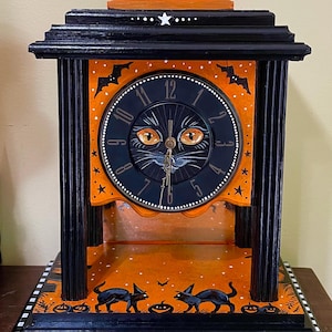 May include: A decorative mantel clock featuring a black cat face on the clock dial with orange eyes. The frame is black and orange with painted bats, stars, and cats. It has a white and black checkered base and a white handle on the top.