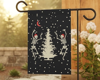 May include: A black garden flag with a white Christmas tree and two skeletons wearing Santa hats dancing. The flag has a white crescent moon and white snowflakes on a black background.