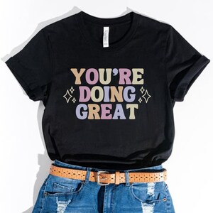 May include: Black t-shirt with the text "YOU'RE DOING GREAT" in colorful letters with sparkling stars on either side of the text. The shirt is paired with denim shorts and a brown belt.