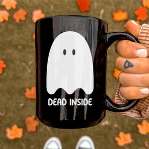 Puede incluir: Taza de cer&aacute;mica negra con un dise&ntilde;o de fantasma blanco y el texto "DEAD INSIDE". La taza se sostiene sobre un fondo de hojas ca&iacute;das. La taza es de tama&ntilde;o est&aacute;ndar para caf&eacute; o t&eacute;.