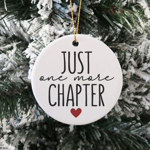 May include: White ceramic Christmas ornament with black text that reads "Just one more CHAPTER" with a small red heart below the text. The ornament is hanging on a snowy artificial Christmas tree.