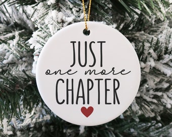 May include: White ceramic Christmas ornament with black text that reads "Just one more CHAPTER" with a small red heart below the text. The ornament is hanging on a snowy artificial Christmas tree.