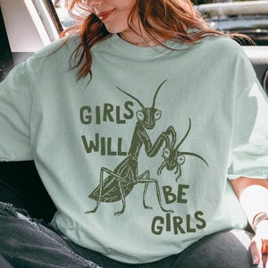 May include: A light green t-shirt with the text "GIRLS WILL BE GIRLS" and a graphic of praying mantises. The person in the image is wearing a brown corduroy hat and sunglasses. The image is taken inside a car.