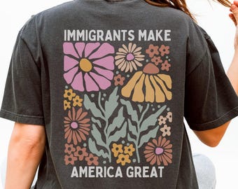 Puede incluir: Camiseta gris oscuro con el texto "IMMIGRANTS MAKE AMERICA GREAT" y un diseño floral en rosa, amarillo y marrón. La camiseta es de corte holgado y tiene mangas cortas.