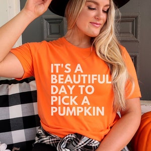 May include: An orange t-shirt with the text "IT'S A BEAUTIFUL DAY TO PICK A PUMPKIN" in white. A black hat is being held. A large orange pumpkin sits on a porch. A black and white checkered pillow and sunflowers are also visible.