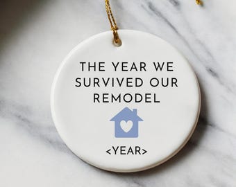May include: A white ceramic ornament with gold string, featuring the text "THE YEAR WE SURVIVED OUR REMODEL" and a blue house graphic. The ornament is surrounded by gold ribbon, wrapping paper, and gold scissors.