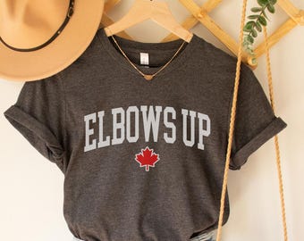 Könnte beinhalten: Ein dunkelgraues V-Ausschnitt-T-Shirt mit dem Text "ELBOWS UP" in Weiß mit einem roten Ahornblatt. Das Shirt hängt an einem Holzgestell mit einer Strohtasche und einem braunen Hut.
