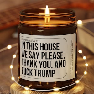May include: A lit brown glass jar candle with a white label that reads "IN THIS HOUSE WE SAY PLEASE, THANK YOU, AND FUCK TRUMP." The candle sits on a small wooden coaster, surrounded by string lights.