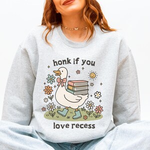 May include: A light gray sweatshirt with a graphic of a white duck wearing blue boots and carrying books, surrounded by flowers and the text "honk if you love recess."