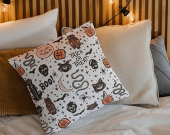 Peut inclure: Un oreiller blanc avec un motif sur le thème d'Halloween représentant des chats noirs, des citrouilles, des chauves-souris, des serpents, des crânes et le texte "Trick or Treat" et "Boo!" L'oreiller est sur un lit avec des draps blancs et un oreiller beige.
