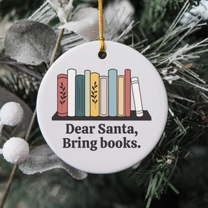 May include: Round white ceramic Christmas ornament with a gold string. The ornament features a bookshelf with colorful books and the text "Dear Santa, Bring books."