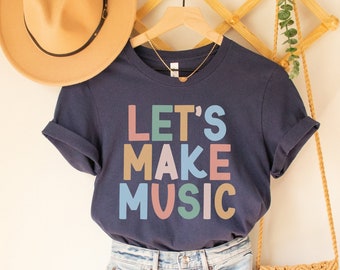 Pode incluir: Uma camiseta azul marinho com o texto "LET'S MAKE MUSIC" em letras coloridas. A camiseta está pendurada em um cabide de madeira com um chapéu de palha e uma bolsa de tecido.