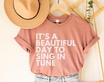 May include: A pink t-shirt with the text "It's a beautiful day to sing in tune" in white lettering. The shirt is paired with denim shorts and a straw hat.