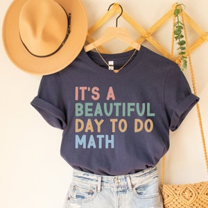 May include: A navy blue t-shirt with the text "It's a beautiful day to do math" in rainbow colors. The shirt is hanging on a wooden rack with a straw hat and a woven bag.