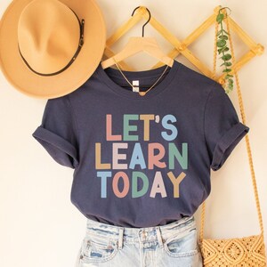 May include: A navy blue t-shirt with the text "LET'S LEARN TODAY" in colorful block letters. The shirt is paired with denim shorts, a tan hat, and a macrame crossbody bag. A gold necklace hangs on the shirt.