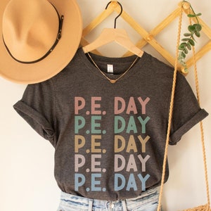 May include: A dark gray t-shirt with the text "P.E. DAY" in multiple pastel colors. The shirt is paired with light wash denim shorts, a tan hat, and a woven crossbody bag. A wooden hanger and decorative wall piece are also visible.