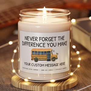 May include: A lit soy candle in a glass jar with a label that reads "NEVER FORGET THE DIFFERENCE YOU MAKE" and a school bus graphic. The candle sits on a wooden coaster, with string lights and a guitar in the background.