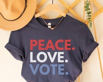 Pode incluir: Camiseta azul marinho com as palavras "PEACE. LOVE. VOTE." em vermelho e azul. A camiseta é combinada com shorts jeans, um chapéu bege e uma bolsa crossbody tecida. Um colar de ouro pende da camiseta.