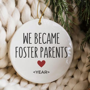 May include: A white ceramic ornament with the text "WE BECAME FOSTER PARENTS" in black, a red heart, and the word "<YEAR>" below. The ornament hangs from a gold string and is set against a white knitted blanket and greenery.