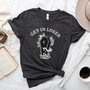 May include: A dark grey t-shirt with the text "GET IN LOSER" printed in white. The shirt features a black and white illustration of a coffin with a crescent moon and flowers.