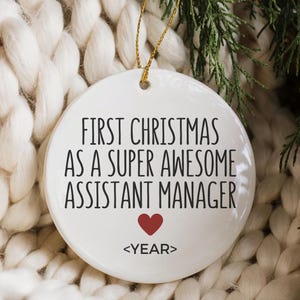 May include: A white ceramic Christmas bauble with gold string, inscribed with "FIRST CHRISTMAS AS A SUPER AWESOME ASSISTANT MANAGER" and a red heart. The bauble is set against a white knitted background and greenery.