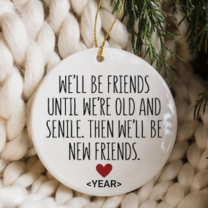 May include: A white ceramic ornament with a gold string for hanging. The ornament has the text "WE'LL BE FRIENDS UNTIL WE'RE OLD AND SENILE. THEN WE'LL BE NEW FRIENDS." with a red heart and the word "<YEAR>" below. The ornament is set against a white knitted background.