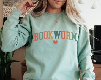 May include: A light blue sweatshirt with the text "BOOKWORM" in colorful letters and a small red heart. The sweatshirt is worn by a person sitting down.