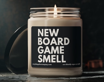 May include: A beige candle with a black label that reads "NEW BOARD GAME SMELL" and the website "everythingafterparty.com". The candle is lit and the flame is visible. The candle is on a dark gray surface.