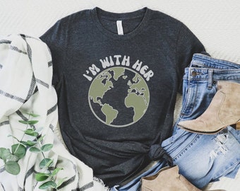 Può includere: Una maglietta grigio scuro con il testo "I'm with her" in un font retrò, con un grafico di globo terrestre verde e grigio. La maglietta è abbinata a jeans blu e stivaletti marroni.