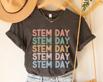 May include: A dark gray t-shirt with the text "STEM DAY" repeated five times in different pastel colors. The shirt is hanging on a wooden hanger with a straw hat and a woven bag.