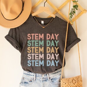 May include: A dark grey t-shirt with the text "STEM DAY" repeated five times in different pastel colours. The shirt is hanging on a wooden hanger with a straw hat and a woven bag.