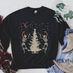 May include: Black sweatshirt with a Christmas tree and two skeletons wearing Santa hats dancing around it. The skeletons are white and the tree is beige. There is a crescent moon and falling snow in the background.