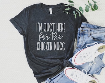 May include: A dark grey t-shirt with the text "I'm just here for the chicken nugs" in white lettering. The shirt is laid out on a white surface with a pair of blue jeans and white trainers.