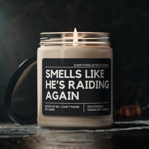 May include: A beige candle with a black label that reads "Smells Like He's Raiding Again". The label also includes the text "Everything After Company", "Notes of No, I Can't Pause My Game", and "Eco-Friendly Vegan Soy Candle".