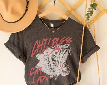 May include: A dark gray t-shirt with a graphic of a roaring cat and the text "Childless Cat Lady". The shirt is paired with light blue denim shorts and a tan crocheted bag.