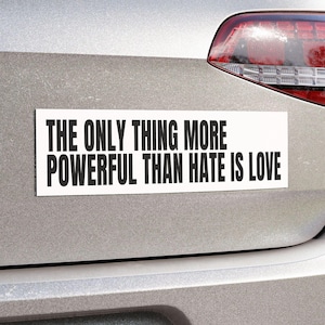 May include: A white rectangular bumper sticker with black text that reads "THE ONLY THING MORE POWERFUL THAN HATE IS LOVE" is attached to the rear of a silver car. The car's taillight is visible.