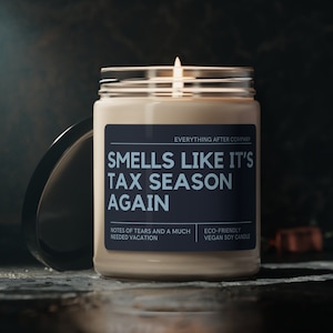 May include: A beige candle with a black label that reads "Smells Like It's Tax Season Again". The label also includes the text "Everything After Company", "Notes of tears and a much needed vacation", and "Eco-friendly vegan soy candle".