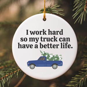 May include: A white ceramic Christmas ornament with a blue pickup truck carrying a Christmas tree. The ornament has the text "I work hard so my truck can have a better life."