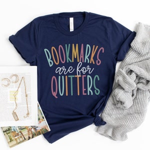May include: Navy blue t-shirt with the text "BOOKMARKS are for QUITTERS" in colorful letters. A gray sweater and a book with jewelry are next to the shirt. The shirt is a casual wear item.