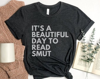 Puede incluir: Una camiseta gris oscuro con el texto "It's a beautiful day to read smut" impreso en blanco en la parte delantera. La camiseta está sobre una superficie blanca con un suéter marrón, una taza blanca, un libro y hojas verdes.