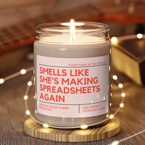 May include: A brown glass candle with a white label that reads "Smells like she's making spreadsheets again". The label also includes the text "Everything After Company", "Notes of color coded perfection", and "Eco-friendly vegan soy candle".