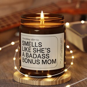 Può includere: Un barattolo di candela in vetro marrone acceso con il testo "Smells Like She's A Badass Bonus Mom" su un'etichetta bianca. La candela è posizionata su un piccolo sottobicchiere di legno, con una stringa di luci e una chitarra sullo sfondo.