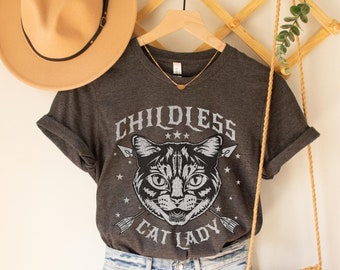 May include: A dark gray t-shirt with a cat graphic and the text "Childless Cat Lady". The shirt is on a hanger with a straw hat and a woven bag.