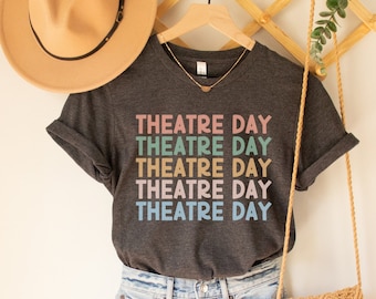 May include: A dark gray t-shirt with the text "THEATRE DAY" repeated five times in different pastel colors. The shirt is hanging on a wooden hanger with a straw hat and a woven bag.