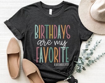 May include: A dark gray t-shirt with the text "Birthdays are my favorite" in colorful letters. The text is in a playful font and is centered on the shirt. The shirt is laid flat on a white surface with a hat, shoes, and a sprig of greenery.