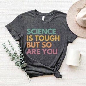 May include: A dark gray t-shirt with the text "SCIENCE IS TOUGH BUT SO ARE YOU" printed in colorful letters. The letters are in a gradient of green, orange, yellow, and pink.